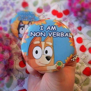 May include: A round blue button with the text "I AM NON VERBAL" and a cartoon dog illustration. The button is held in a hand. A second button with the text "I HAVE AN INVISIBLE DISABILITY" is visible in the background.