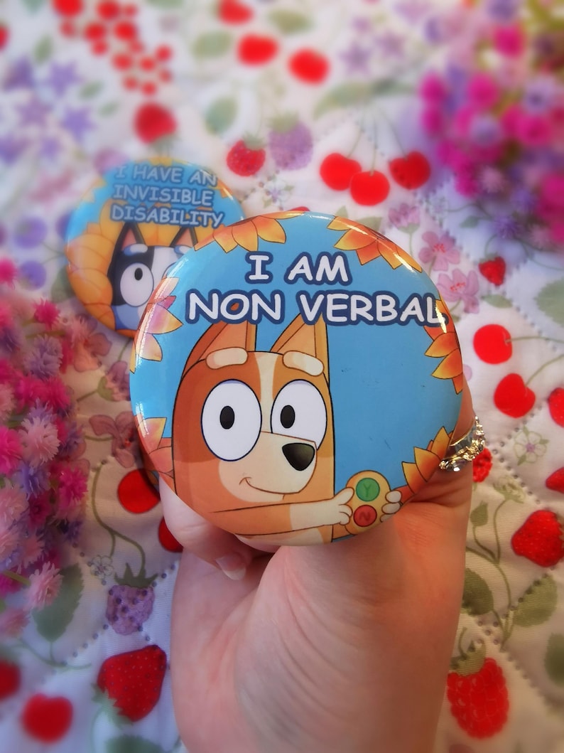 May include: A round blue button with the text "I AM NON VERBAL" and a cartoon dog illustration. The button is held in a hand. A second button with the text "I HAVE AN INVISIBLE DISABILITY" is visible in the background.
