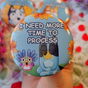 May include: A collection of round, colorful buttons with cartoon characters and text. One button reads "I NEED MORE TIME TO PROCESS" with a blue dog character. Other buttons have text like "I HAVE AN INVISIBLE DISABILITY".