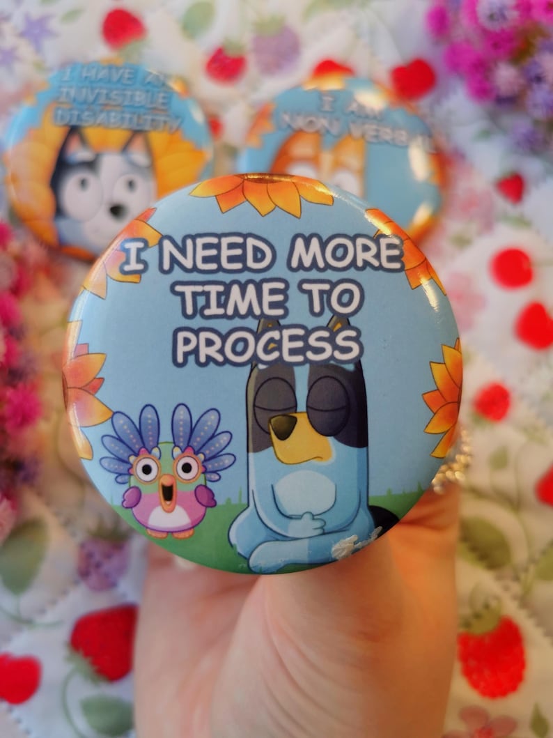 May include: A collection of round, colorful buttons with cartoon characters and text. One button reads "I NEED MORE TIME TO PROCESS" with a blue dog character. Other buttons have text like "I HAVE AN INVISIBLE DISABILITY".
