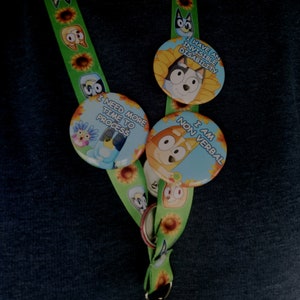 May include: Green lanyard with cartoon character and sunflower pattern. Attached are three round buttons with cartoon characters and text: "I HAVE AN INVISIBLE DISABILITY", "I NEED MORE TIME TO PROCESS", and "I AM NON VERBAL".
