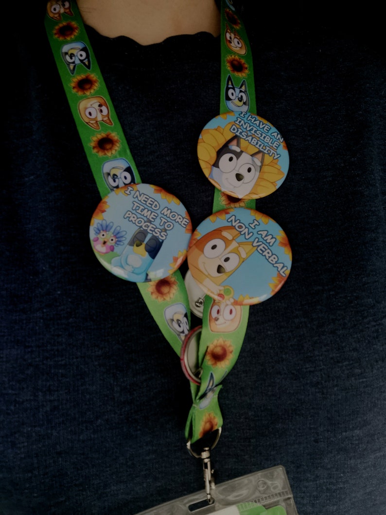 May include: Green lanyard with cartoon character and sunflower pattern. Attached are three round buttons with cartoon characters and text: "I HAVE AN INVISIBLE DISABILITY", "I NEED MORE TIME TO PROCESS", and "I AM NON VERBAL".