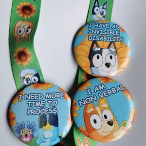 May include: Three round buttons with cartoon characters and text. The buttons read "I HAVE AN INVISIBLE DISABILITY", "I NEED MORE TIME TO PROCESS", and "I AM NON VERBAL". The buttons are on a green lanyard with cartoon characters and sunflowers.
