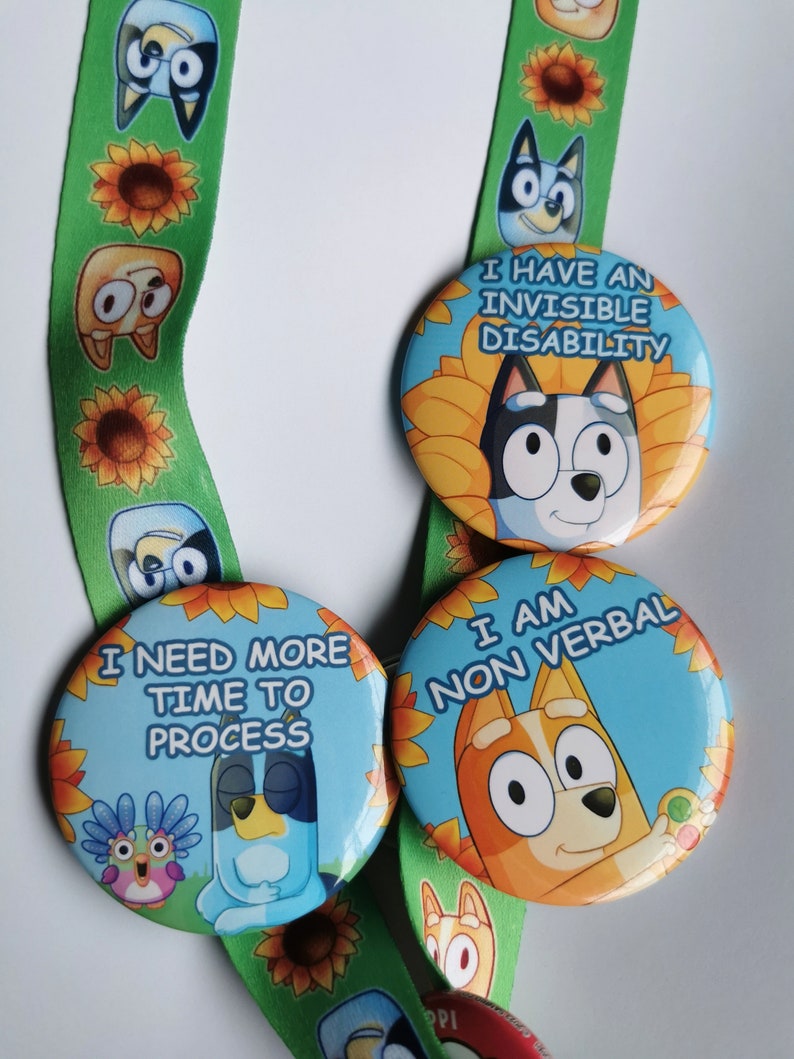 May include: Three round buttons with cartoon characters and text. The buttons read "I HAVE AN INVISIBLE DISABILITY", "I NEED MORE TIME TO PROCESS", and "I AM NON VERBAL". The buttons are on a green lanyard with cartoon characters and sunflowers.