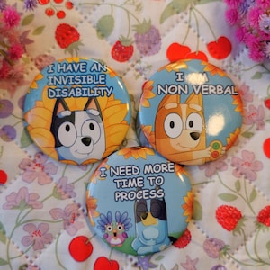 May include: Three round buttons with cartoon dog illustrations and text. The buttons have blue, yellow, and white designs. The text on the buttons reads: "I HAVE AN INVISIBLE DISABILITY", "I AM NON VERBAL", and "I NEED MORE TIME TO PROCESS".