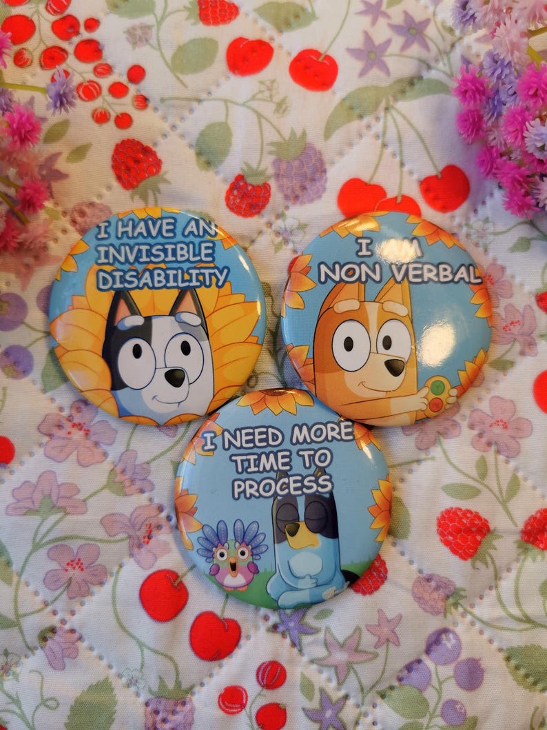 May include: Three round buttons with cartoon dog illustrations and text. The buttons have blue, yellow, and white designs. The text on the buttons reads: "I HAVE AN INVISIBLE DISABILITY", "I AM NON VERBAL", and "I NEED MORE TIME TO PROCESS".