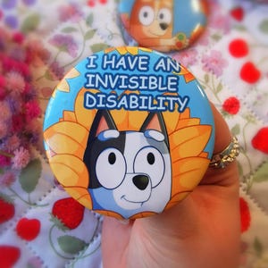 May include: A round button with the text "I HAVE AN INVISIBLE DISABILITY" in blue and white. The button features a cartoon dog character with a blue and white face, surrounded by yellow sunflower petals. Another button is visible in the background.