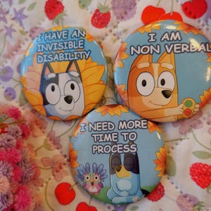 May include: Three round pinback buttons with cartoon dog illustrations and text. The buttons have blue, yellow, and orange backgrounds. Text includes "I HAVE AN INVISIBLE DISABILITY", "I AM NON VERBAL", and "I NEED MORE TIME TO PROCESS".