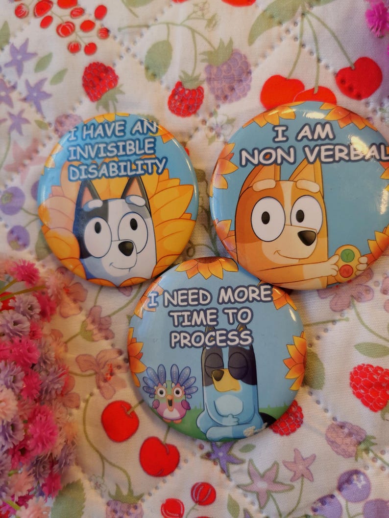 May include: Three round pinback buttons with cartoon dog illustrations and text. The buttons have blue, yellow, and orange backgrounds. Text includes "I HAVE AN INVISIBLE DISABILITY", "I AM NON VERBAL", and "I NEED MORE TIME TO PROCESS".