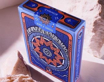 Könnte beinhalten: Ein blaues und oranges Spielkartenspiel mit einem Sternburst-Design und dem Text "CYGNUS 590/1000" auf der Vorderseite. Das Deck heißt "Celestial Planes".