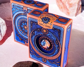 Könnte beinhalten: Zwei orangefarbene und blaue Spielkartenspiele mit einem himmlischen Design. Das obere Deck hat die Nummer 130/1000 und das Wort "OMEGA" aufgedruckt.