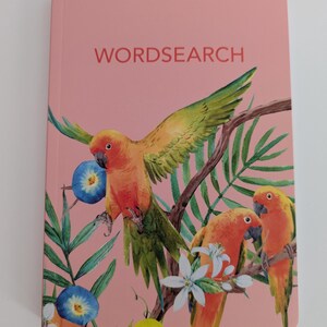 May include: A pink wordsearch book featuring a watercolor illustration of three orange and green parrots perched on branches among green palm leaves, white flowers, and blue morning glory blossoms with the text WORDSEARCH printed in red.