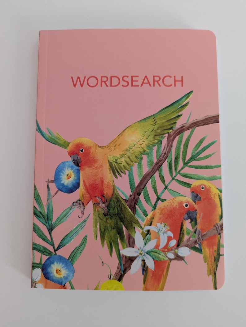 May include: A pink wordsearch book featuring a watercolor illustration of three orange and green parrots perched on branches among green palm leaves, white flowers, and blue morning glory blossoms with the text WORDSEARCH printed in red.