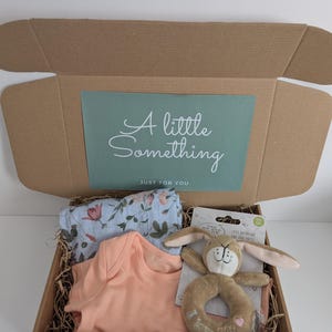 May include: A brown cardboard gift box containing a light blue floral patterned swaddle, a solid peach-colored baby bodysuit, and a tan plush rabbit ring rattle with the text I love you. The box lid features a green insert with the text A little Something JUST FOR YOU.
