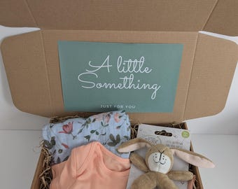 May include: A brown cardboard gift box containing a light blue floral patterned swaddle, a solid peach-colored baby bodysuit, and a tan plush rabbit ring rattle with the text I love you. The box lid features a green insert with the text A little Something JUST FOR YOU.