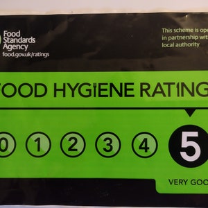 May include: A rectangular Food Standards Agency food hygiene rating sticker with a black background and a large green central panel. It displays the text FOOD HYGIENE RATING, a scale from 0 to 5, with the number 5 highlighted and labeled VERY GOOD.