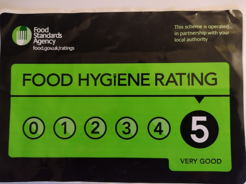May include: A rectangular Food Standards Agency food hygiene rating sticker with a black background and a large green central panel. It displays the text FOOD HYGIENE RATING, a scale from 0 to 5, with the number 5 highlighted and labeled VERY GOOD.