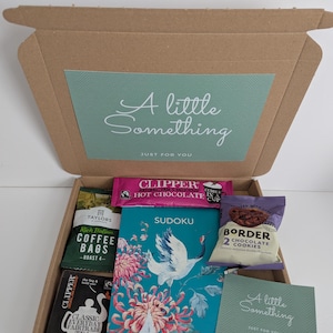 May include: A brown cardboard gift box containing a Sudoku book with a crane and flower illustration, a packet of Clipper hot chocolate, Taylors coffee bags, Clipper tea bags, a pack of Border chocolate cookies, and a small card labeled A little Something.