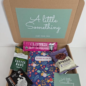 May include: A cardboard gift box containing a word search book with a floral pattern, a pink Clipper hot chocolate sachet, a green Taylors coffee bag, a black Clipper tea bag, a pack of Border chocolate cookies, and a card reading A little Something.