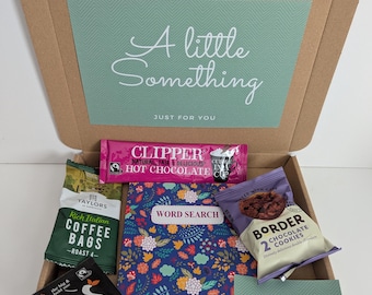 May include: A cardboard gift box containing a word search book with a floral pattern, a pink Clipper hot chocolate sachet, a green Taylors coffee bag, a black Clipper tea bag, a pack of Border chocolate cookies, and a card reading A little Something.