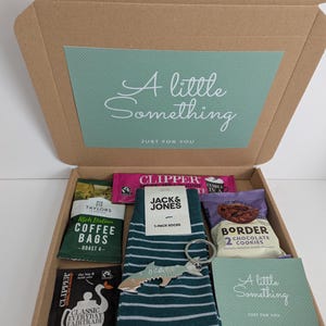 May include: A cardboard gift box containing a pair of dark green striped socks with a Jack & Jones label, a silver fish-shaped keychain, a pack of Taylors coffee bags, two Clipper tea bags, a pack of Border chocolate cookies, and two cards.