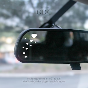 May include: A car's rearview mirror with a series of white heart decals. The decals are arranged in a cascading pattern. The text "GDC Design Co." is visible in the upper right corner. The text "Decals pictured here are NOT to size. View description for proper sizing information." is at the bottom.