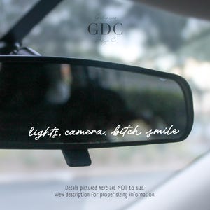 May include: A black car rearview mirror with the words "lights, camera, bitch smile" in white script. The image includes the GDC Design Co. logo and a disclaimer about decal sizing.