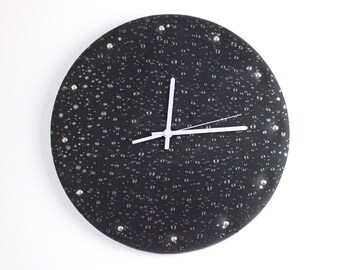 Puede incluir: Un reloj redondo negro con agujas blancas y detalles plateados. La esfera del reloj tiene un diseño texturizado que se asemeja a gotas de lluvia.