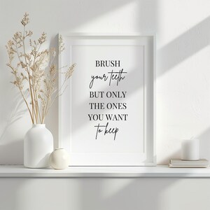 May include: A white framed print with the text "BRUSH your teeth BUT ONLY THE ONES YOU WANT to keep" in black script. A white vase with dried flowers and a small white vase are on the shelf. A white candle and books are on the right.