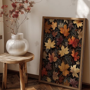 Op de afbeelding: Een ingelijst kunstwerk met een patroon van herfst esdoornbladeren in rood, oranje en geel tegen een donkere achtergrond. Een witte vaas met gedroogde bloemen staat op een houten bijzettafel.