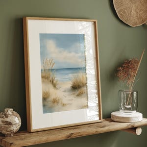 Op de afbeelding: Een ingelijste aquarelprint van een strandscène. Het kunstwerk toont een zanderig pad dat naar de oceaan leidt onder een bewolkte blauwe lucht. Hoge, gedroogde grassen omlijsten het pad. De lijst is van lichtgekleurd hout en wordt tentoongesteld op een houten plank.