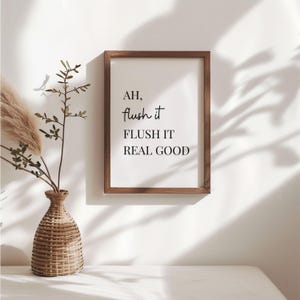 May include: A framed print with the text "AH, flush it, FLUSH IT REAL GOOD" in black script. The frame is a dark wood colour. A decorative vase with dried plants sits to the left of the frame.