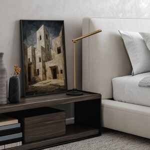 May include: A bedroom scene with a dark wood nightstand displaying a framed abstract art print, a black vase with dried flowers, and a gold desk lamp. A bed with a white headboard and bedding is in the background.