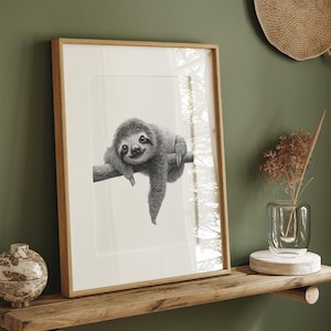 May include: A framed monochrome print of a sloth on a branch. The sloth is depicted with a smiling expression. The frame is light wood. The print is displayed on a wooden shelf, alongside decorative items, against a green wall.
