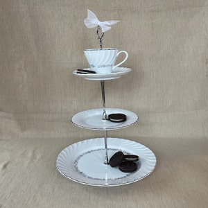 May include: A three-tiered serving stand with a teacup, saucer, and cookies. The white porcelain plates have a silver trim and a floral design. A white bow adorns the top. The stand is made of metal.