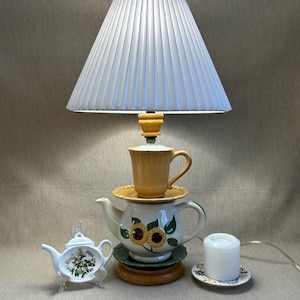 May include: A unique lamp made from a teapot, teacup, and saucer base, topped with a pleated lampshade. The teapot features sunflower designs. A small white teapot and a candle on a saucer are also present.