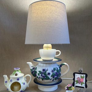 May include: A unique lamp made from a teapot, teacup, and saucer, with a lampshade. The teapot is decorated with purple flowers. A small teapot with a candle inside and a framed cross-stitch are also present.