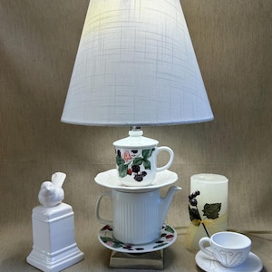 May include: A unique lamp made from a white teapot, teacup, and saucer, topped with a white lampshade. The teacup and saucer have a floral design with berries. A white bird statue and a candle with berry accents are also present.