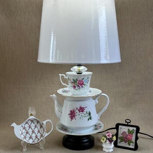 May include: A unique lamp made from a teapot, teacup, and saucer, with a white lampshade. The teapot and teacup are decorated with floral patterns. A small framed cross-stitch and a miniature vase with flowers are also present.