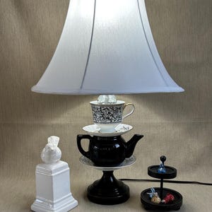 May include: A unique lamp with a white lampshade, a black teapot base, and a teacup with a black and white design. A white bird statue and a small black tiered tray with candies are also present. The lamp is on a beige surface.