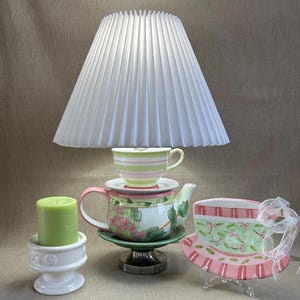 May include: A white pleated lampshade sits above a teacup and teapot. A green candle sits in a white candle holder. A decorative teacup-shaped plate with pink and green accents is also present. The teapot and teacup have floral designs.