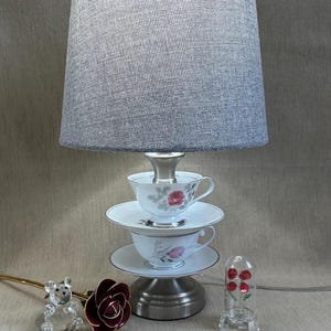 May include: A unique table lamp with a gray fabric shade, supported by a base made of stacked teacups and saucers. The teacups feature floral designs. Decorative glass rose and bear figurines are also present.