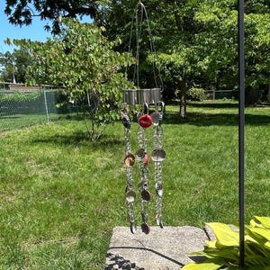 May include: A handmade wind chime with a metal frame and chains. The chains are adorned with bottle caps and metal discs. The wind chime is hanging from a black metal hook in a garden setting.