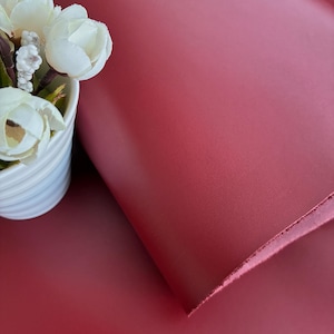 May include: A close-up of a sheet of red leather with a smooth texture. The leather is partially rolled, revealing its thickness and the raw edge. A white ceramic pot with white flowers is in the upper left corner.