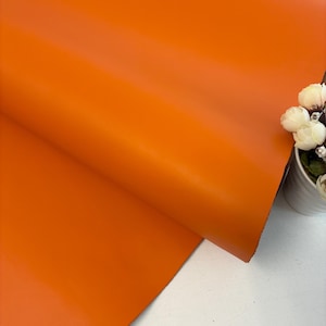 Puede incluir: Primer plano de una vibrante lámina de cuero naranja, doblada para revelar su textura suave. El color es un tono rico y sólido, con sutiles variaciones de tono. El cuero se utiliza probablemente para proyectos de artesanía o tapicería.