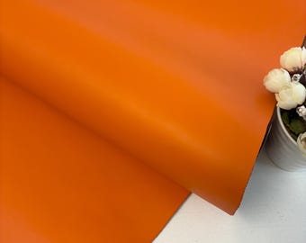 May include: Close-up of a vibrant orange leather sheet, folded to reveal its smooth texture. The color is a rich, solid hue, with subtle variations in tone. The leather is likely used for crafting or upholstery projects.