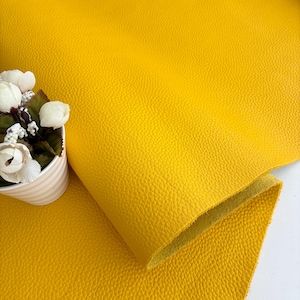 May include: A sheet of bright yellow faux leather with a textured surface. The material is folded over, revealing a lighter yellow underside. A small white ceramic pot with white flowers and green leaves sits to the left of the leather.
