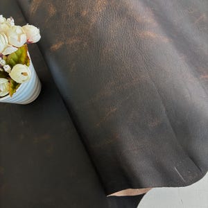 May include: A large piece of dark brown leather with a distressed, textured surface showing lighter tan highlights. Beside the leather is a white fluted container holding a cluster of small, cream-coloured artificial flowers with green leaves.
