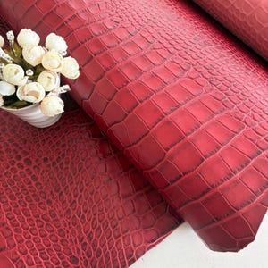 Peut inclure: Une feuille de cuir rouge présentant une texture en relief façon peau de crocodile avec des ombres sombres dans les rainures. Un petit vase blanc contenant un bouquet de fleurs artificielles blanches est placé sur le côté gauche.