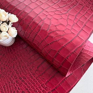 May include: A sheet of deep red leather featuring a prominent embossed crocodile skin texture with dark shading in the crevices. The material is folded over to show the underside and is positioned next to a small white vase with white flowers.